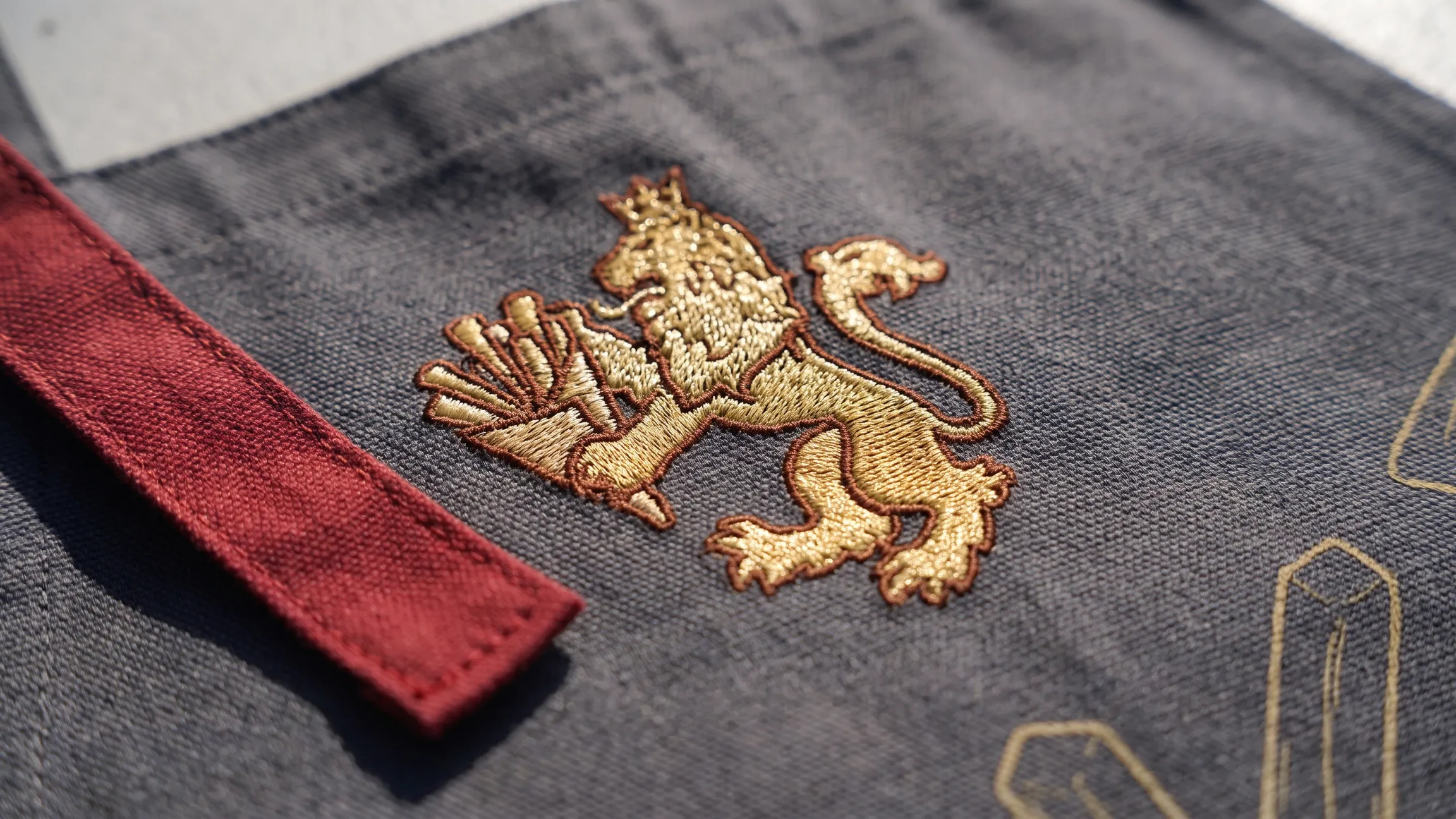 Gold Friteur lion crest embroidered on a grey apron with an oxblood strap