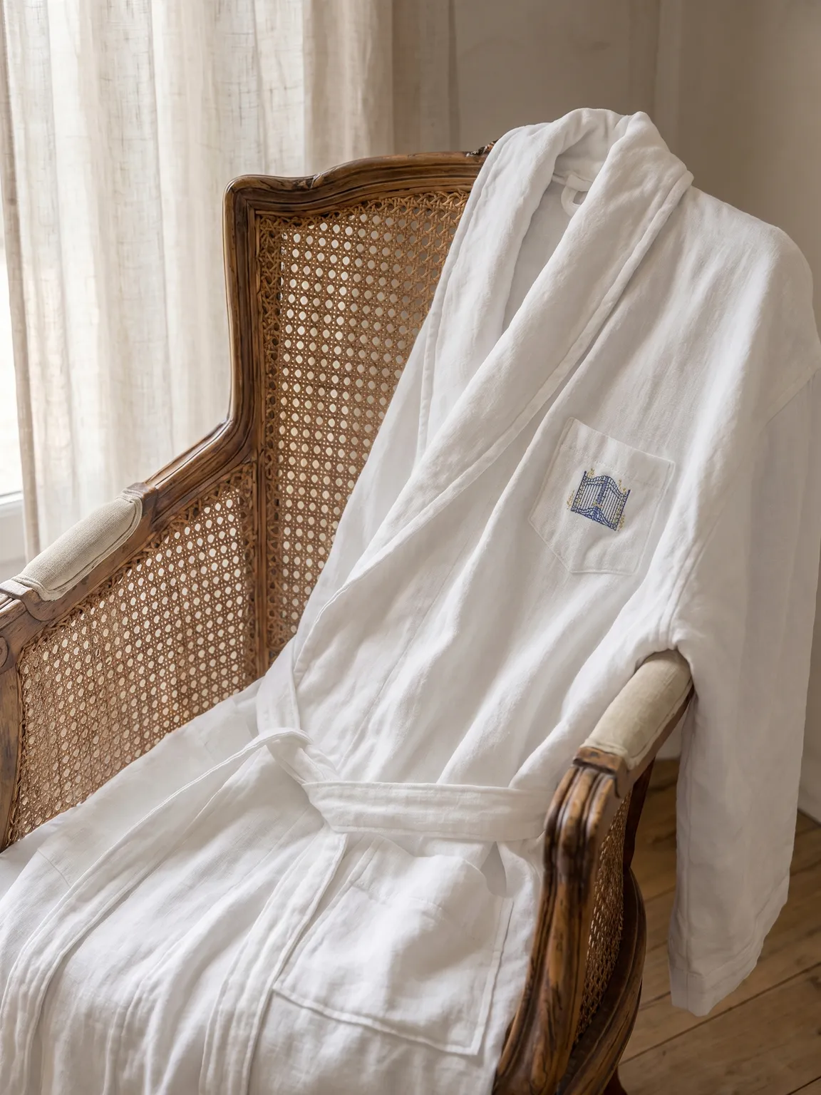 White linen robe embroidered with the La Belle Échappée crest, draped over a cane chair