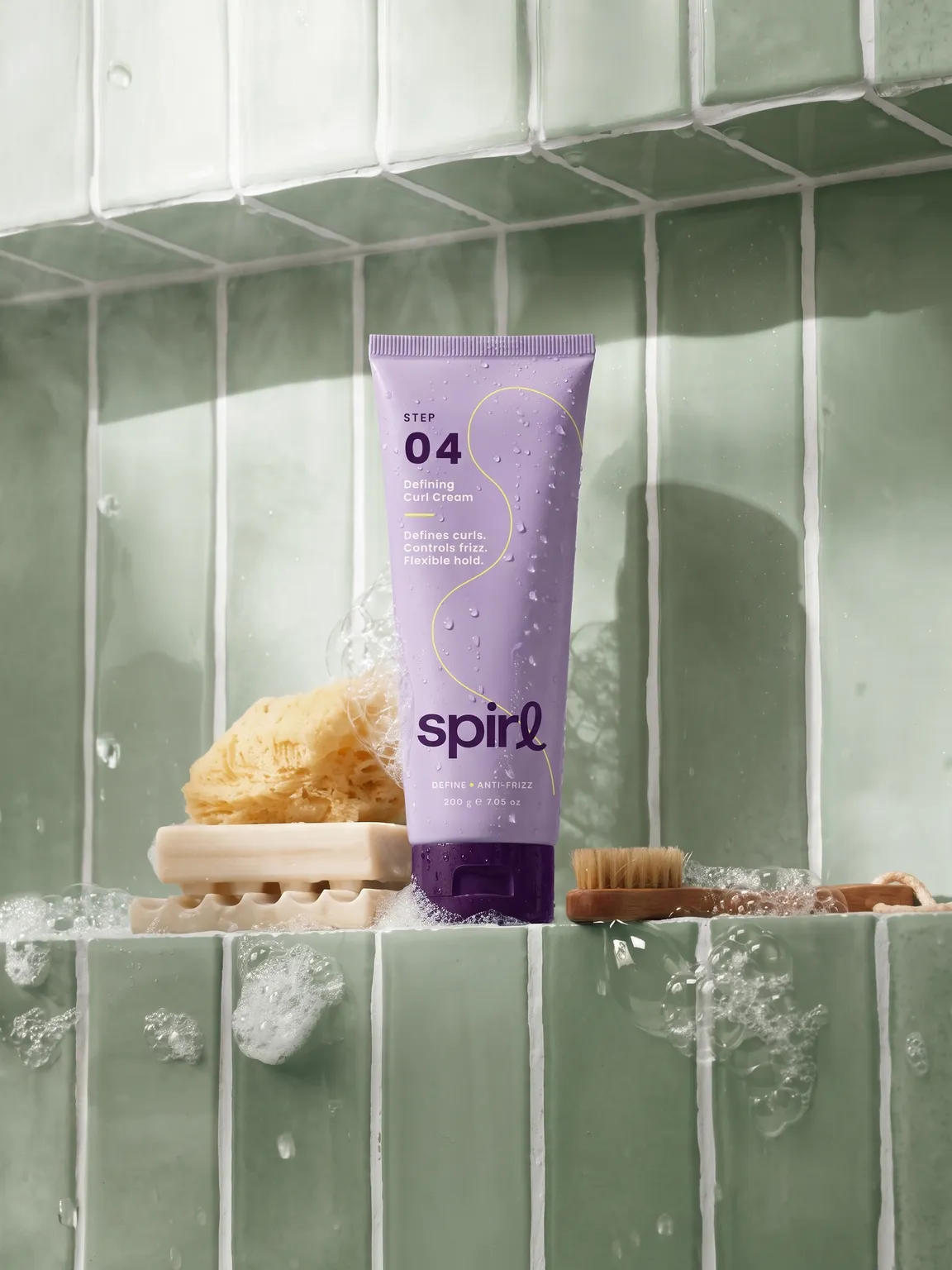 Lilac Step 04 Defining Curl Cream tube on a sage-tiled shower shelf with sponge, soap and brush
