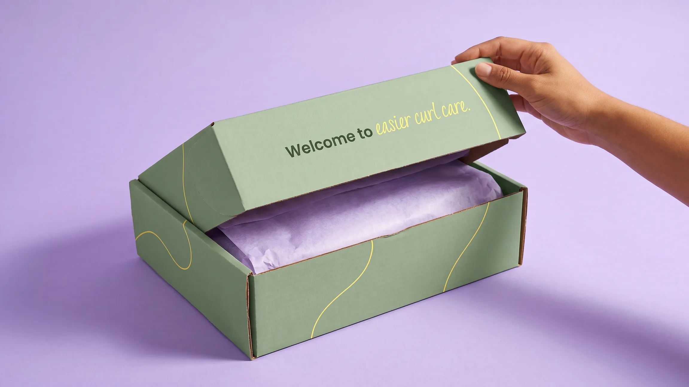 Hand lifting the lid of the sage-green Spirl shipper box, printed "Welcome to easier curl care"
