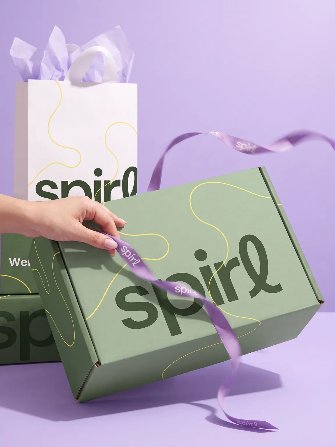 Hand pulling a lilac Spirl ribbon around the sage-green shipper box