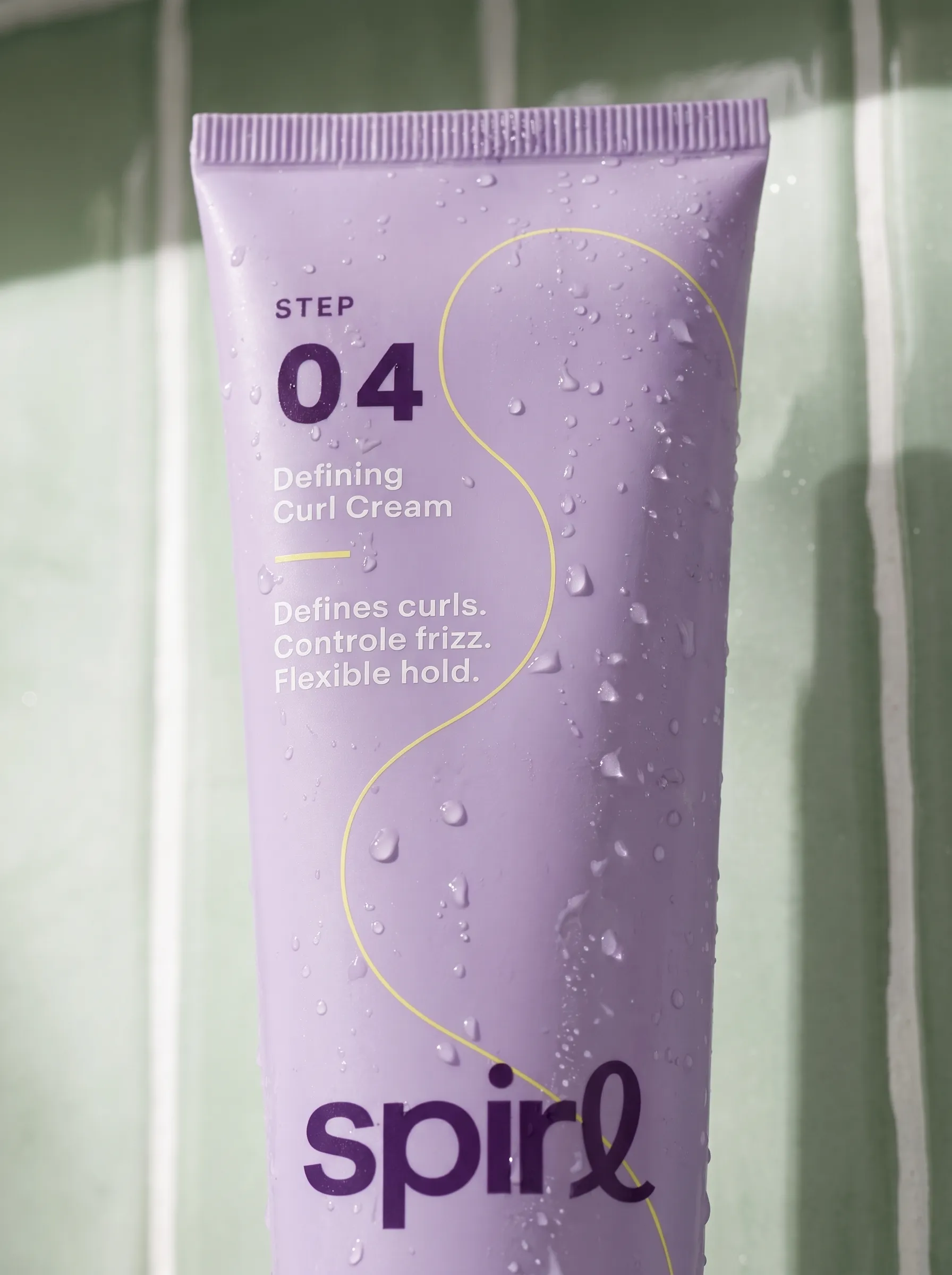 Water beading on the lilac Step 04 Defining Curl Cream tube against green shower tiles