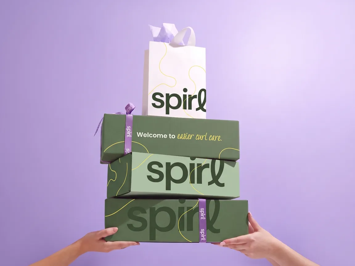 Hands holding a stack of Spirl shipper boxes and a gift bag reading “Welcome to easier curl care” on a lavender background