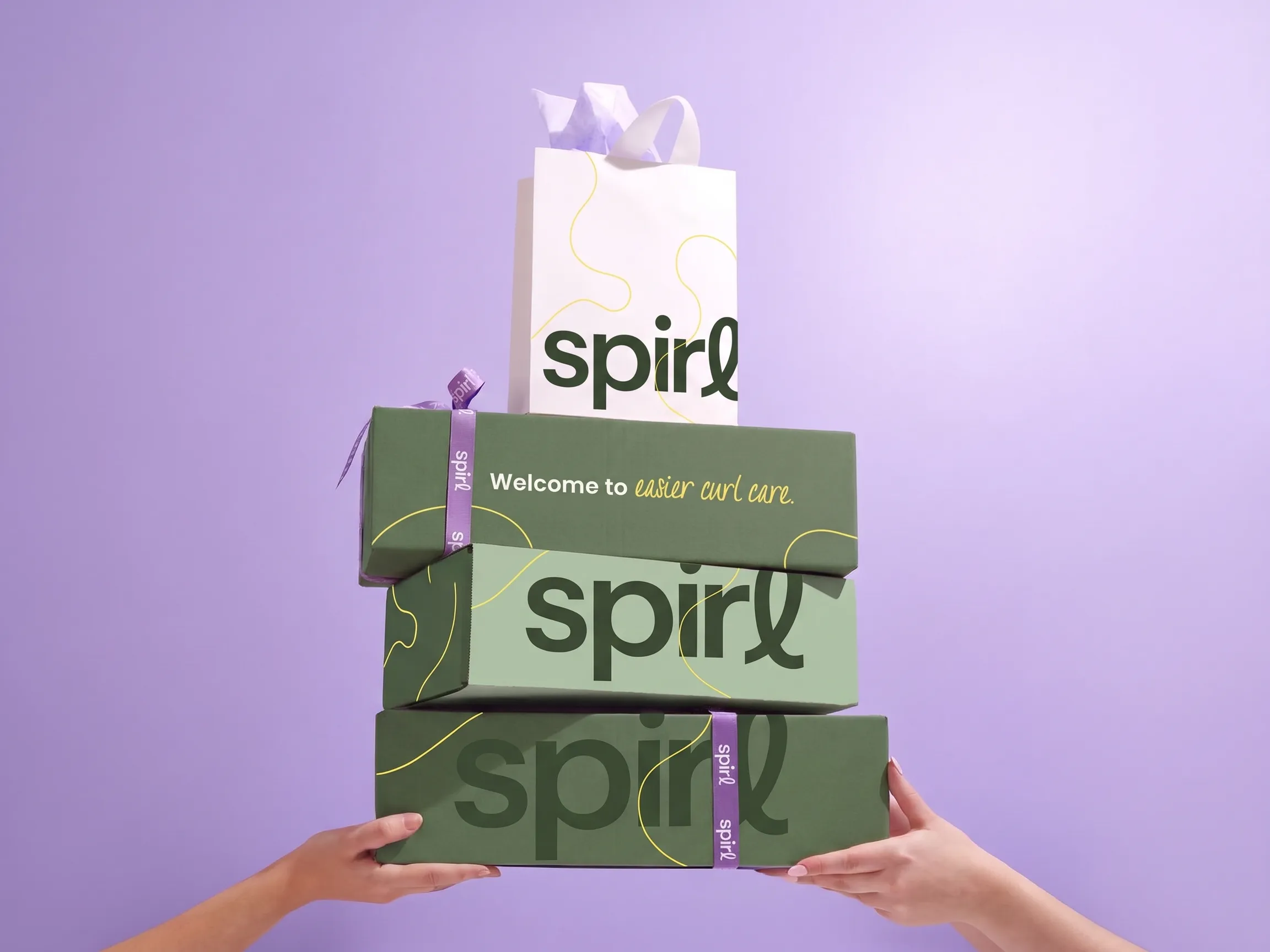 Hands holding a stack of Spirl shipper boxes and a gift bag reading “Welcome to easier curl care” on a lavender background